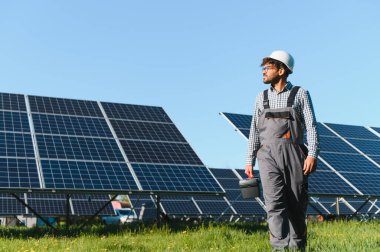 Fotovoltaik panellerin yanında yürüyen orta doğu mühendisi büyük bir güneş çiftliğinde bir alet çantası taşıyor, tulum ve şapka takıyor.
