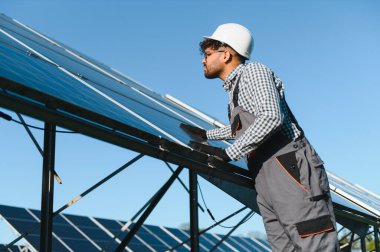 Açık mavi gökyüzü olan güneşli bir günde, büyük bir güneş enerjisi santraline fotovoltaik paneller kurulumu üzerinde çalışan Ortadoğu mühendisi