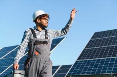 Bir elektrik santraline güneş panelleri yerleştirirken beyaz miğfer ve alet çantasıyla gülümseyen mühendis.