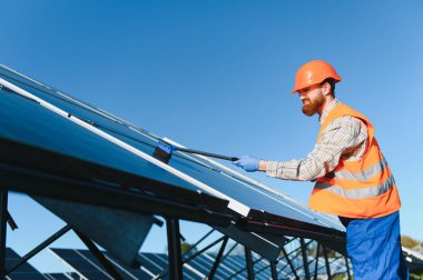 Erkek işçi, açık mavi gökyüzü altında sürdürülebilir ve yeşil enerji çözümlerini teşvik ederek, güneş panelinin kurulumu ve bakımıyla ustalıkla ilgileniyor.