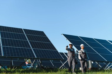 Parlak, güneşli bir gökyüzünün altındaki bir güneş enerjisi santralinde fotovoltaik panellerin kurulmasında işbirliği yapan iki mühendis.