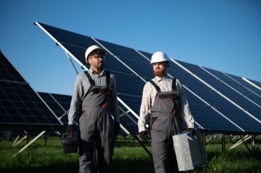 Güneşli bir günde güneş paneli çiftliğinde yürüyen ve aletleri taşıyan iki çoklu etnik mühendis.