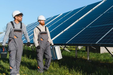 Güneş paneli çiftliğinde yürüyen iki teknisyen, açık bir günde aletleri taşıyor ve güneşin altında birlikte çalışıyorlar.