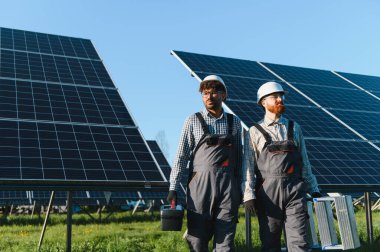 Güneş enerjisi istasyonunda yürüyen iki mühendis, araçlar ve merdiven taşıyorlar, yenilenebilir enerji çözümleri üzerinde birlikte çalışıyorlar.