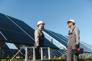 İki mühendis, büyük bir fotovoltaik enerji santralindeki güneş panellerinin kurulumu veya bakımı sırasında atılacak adımları tartışıyor ve araçları taşıyor.