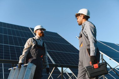 Koruyucu dişli iki işçi, açık mavi gökyüzü altında takım çalışması ve yenilenebilir enerji çözümlerini vurgulayarak, güneş panelleri kurulmasında işbirliği yapıyorlar.