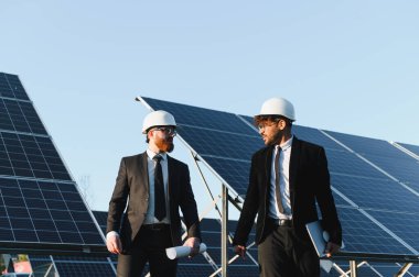 İki mühendis güneş enerjisi istasyonunda güneş panellerinin yanında yürüyorlar ve tartışıyorlar.