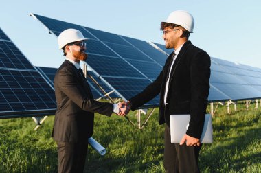 İki mühendis bir güneş enerjisi santralinde el sıkışıyor, fotovoltaik panellerin başarılı kurulumunu kutluyorlar.