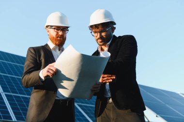 Açık mavi bir gökyüzü altında güneş enerjisi santralini incelerken planlar üzerinde çalışan iki mühendis.