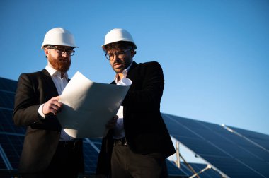 Sert şapkalı iki mühendis güneş paneli çiftliğinin planlarını inceliyor ve yenilenebilir enerji gelişimini tartışıyorlar.