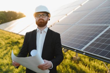 Sert bir şapka takan ve elinde bir planla güneş paneli çiftliğini inceleyen yatırımcı, günbatımında sürdürülebilir enerji girişimlerini vurguluyor.