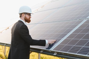 Giysi giyen yatırımcı ve güneş çiftliğindeki bir güneş panelini teftiş ederken, temiz enerji üretimini değerlendirmek için fotovoltaik hücrelere dokunuyor.