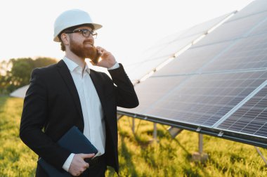 Gülümseyen işadamı elinde tablet ile yeşil alanda güneş panelleri yakınında akıllı telefon kullanıyor.