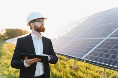 Yatırımcı başlık takıyor ve dijital tablet kullanıyor. Güneş paneli çiftliğini canlı bir günbatımı gökyüzü altında inceliyor. Yenilenebilir enerjiyi vurguluyor.