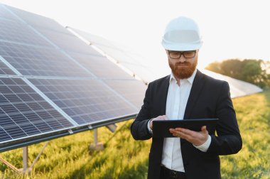 Güneş paneli çiftliğinin gün batımında verimliliğini incelerken dijital tablet kullanan takım elbiseli bir yatırımcı.