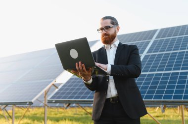 Dizüstü bilgisayar kullanan, yenilenebilir enerji santralindeki güneş panellerini yöneten, sürdürülebilir geleceğe yatırım yapan bir iş adamı.