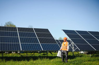 Güneş paneli çiftliğinde yürüyen, alet çantası taşıyan, güvenlik kaskı takan, temiz ve sürdürülebilir enerji üretimine katkıda bulunan mühendis.