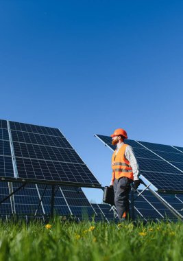 Erkek teknisyen, açık mavi gökyüzünün altındaki enerji santralindeki güneş panellerini incelerken, yürüyen ve alet çantası taşıyor.