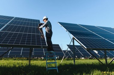 Bir merdiven üzerinde çalışan elektrikçi, büyük bir güneş enerjisi santraline fotovoltaik paneller yerleştiriyor, yenilenebilir enerji üretimine katkıda bulunuyor