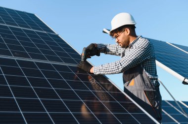 Açık mavi gökyüzü olan güneşli bir günde, bir güneş çiftliğinde çalışan orta doğulu teknisyen tornavida kullanarak fotovoltaik paneller kuruyor.