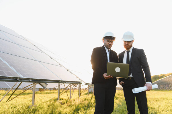 Два инженера в хардхатах анализируют солнечную ферму с ноутбуком, работая над проектом по возобновляемой энергии