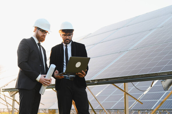 Два инженера в костюмах и касках обсуждают проект с помощью ноутбука на солнечной электростанции