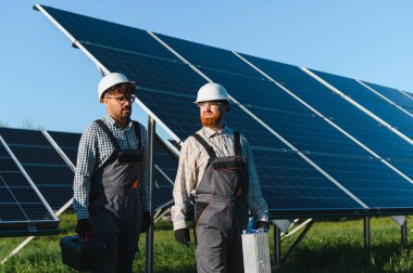 Parlak güneşin altında güneş paneli çiftliğinde yürüyen iki mühendis yenilenebilir enerji çözümleri üzerinde birlikte çalışıyorlar.