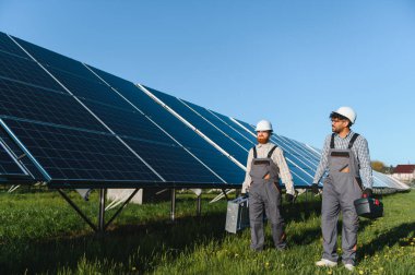 Güneş panelleri yakınlarındaki yeşil bir alanda yürüyen iki mühendis temiz enerjiye ve sürdürülebilir bir geleceğe katkıda bulunuyorlar.