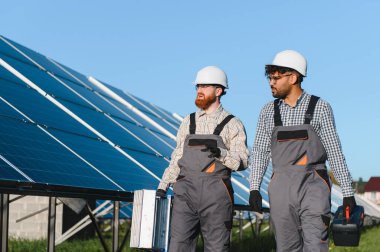 Aletleri taşıyan ve büyük bir güneş paneli çiftliğinde yürüyen iki mühendis bakım ve kurulum hakkında tartışıyorlar.