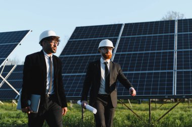 Güneş panelleri alanında yürüyen takım elbiseli iki işadamı yenilenebilir enerji yatırımları ve planlarını tartışıyor.