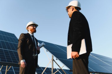 Takım elbiseli ve şapkalı iki mühendis güneş enerjisi istasyonunun planlarını ve laptopunu ellerinde tutmayı tartışıyor.