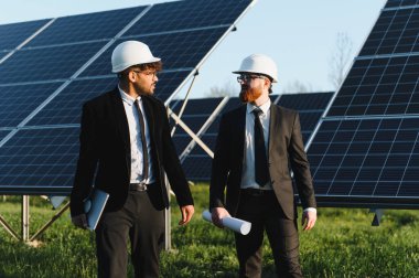 İki iş adamı, büyük bir güneş çiftliğinde güneş panellerinin yanında yürüyen ve tartışan takım elbiseli ve kasklı adamlar.