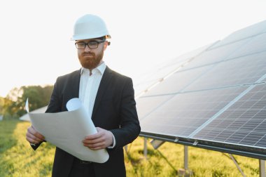 Kasketli bir yatırımcı, güneş paneli çiftliğinin önünde dururken planları inceliyor, yenilenebilir enerji geliştirmeye odaklanıyor.