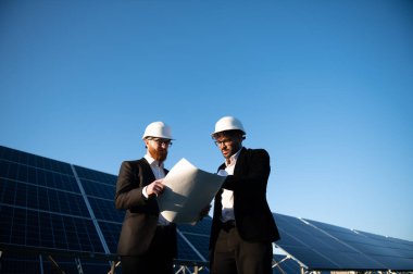 Takım elbiseli iki mühendis, güneş enerjisi santralindeki açık mavi gökyüzünün planlarını inceliyor.