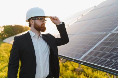 Takım elbiseli bir yatırımcı güneş paneli çiftliğini gün batımında teftiş ederken kaskına dokunuyor.