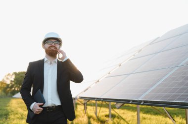 Takım elbiseli ve kasklı yatırımcı cep telefonu kullanıyor ve büyük bir alanda güneş panelleri yakınında tablet tutuyor.