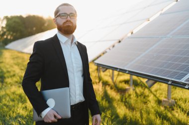 Kendine güvenen işadamı elinde fotovoltaik paneller kullanarak elektrik üreten bir güneş enerjisi santralini teftiş eden dizüstü bilgisayar tutuyor.