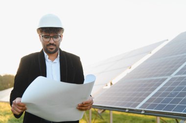 Güneş çiftliğindeki güneş paneli kurulumunu incelerken başlık takan ve elinde plan tutan Arap mühendis.