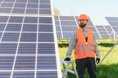 Güvenlik kaskı ve yeleği takan erkek mühendis, bir enerji santralinde güneş panelleri yakınında çalışarak yenilenebilir enerji çözümlerine katkıda bulunuyor.