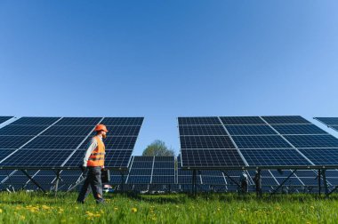 Bir güneş enerjisi istasyonunda güvenlik kaskı takan, yürüyen ve güneş panellerini inceleyen bir mühendis.