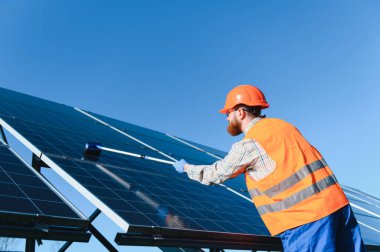 Erkek mühendis fırçayla güneş panellerini temizliyor, açık mavi gökyüzü altında yeşil enerji üretimi için maksimum verimliliği sağlıyor