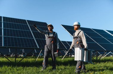 Güneş enerjisi santralinde yürüyen ve çalışan iki mühendis, ellerinde aletlerle güvenlik kaskı takıyorlar.