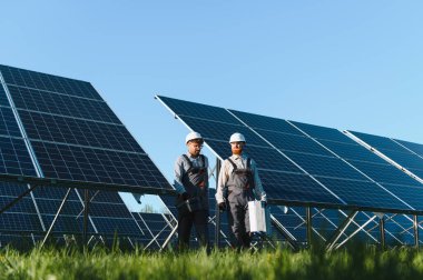 Güneşli bir günde güneş paneli çiftliğinde yürüyen ve araçları taşıyan iki mühendis, yenilenebilir enerji ve sürdürülebilir uygulamalar sergiliyor