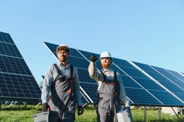 Güneş enerjisi santralinde yürüyen ve çalışan iki mühendis, ellerinde aletlerle ve şapka takıyorlar.