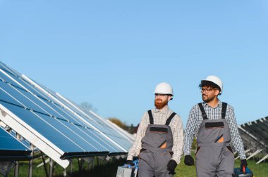 İki çoklu etnik mühendis güneş paneli çiftliğinde yürüyorlar ve aletleri taşıyorlar, sıkı şapkalar ve iş tulumları giyiyorlar.