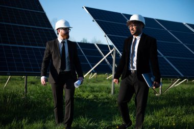 Güneş paneli çiftliğinde yürüyen iki mühendis proje detaylarını tartışıyor ve sürdürülebilir enerji için yenilikçi çözümler üzerinde işbirliği yapıyorlar.