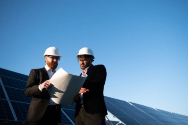 İki mühendis güneş enerjisi santralinin planlarını gözden geçirip yenilenebilir enerji gelişimini tartışıyorlar.