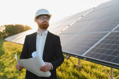 Kask takan ve elinde planlar bulunan yatırımcı güneş paneli çiftliğini gün batımında denetliyor ve yenilenebilir enerji gelişimi ve sürdürülebilirliği vurguluyor.