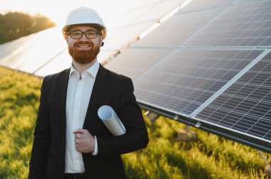 Mühendis elinde planlar tutarken gülümsüyor ve canlı bir günbatımında bir güneş paneli santralini inceliyor, yenilenebilir enerji yeniliğini vurguluyor