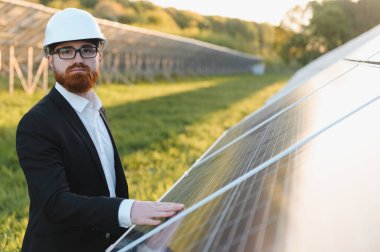 Yeşil bir alanda güneş paneli kurulumunu denetleyen kasklı bir yatırımcı eliyle fotovoltaik hücrelere dokunuyor.